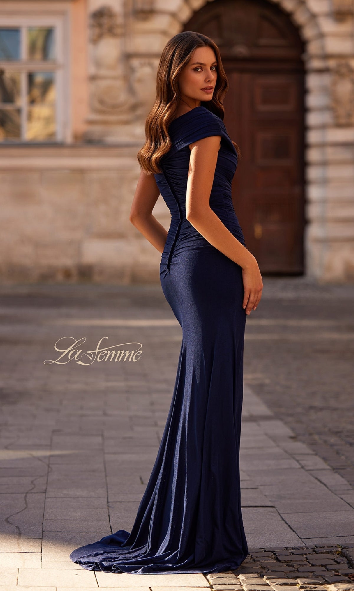 A woman stands outdoors on a cobblestone street, looking over her shoulder in the Cap-Sleeve Long Formal Dress: La Femme 29558.